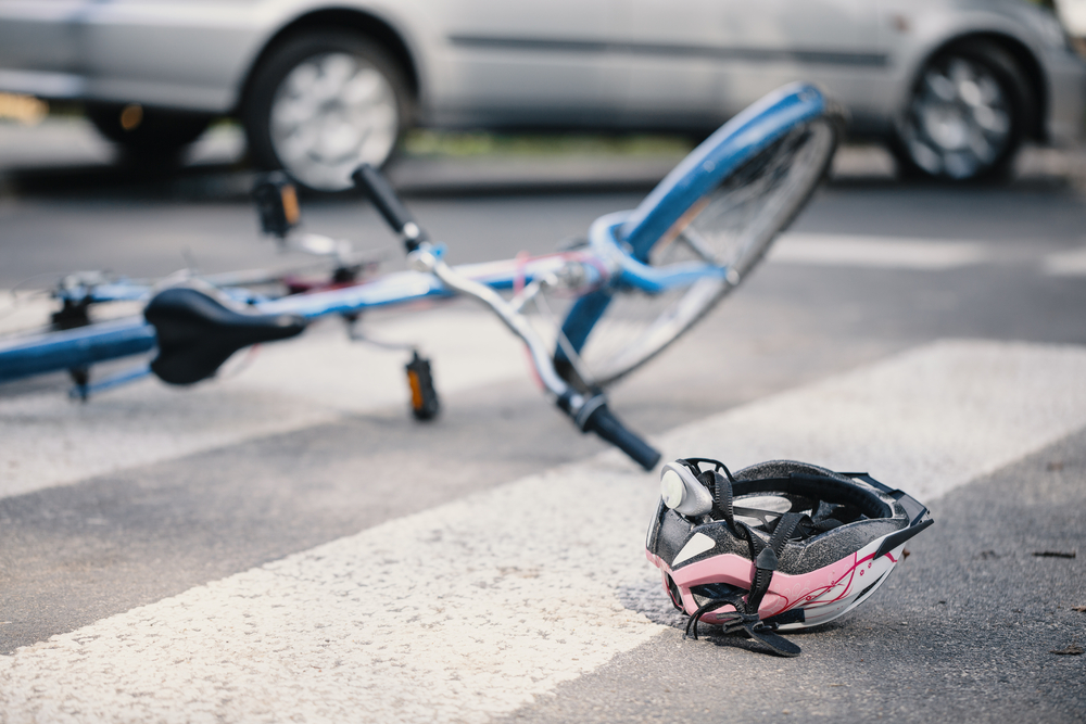 Blue bicycle laying on the ground in a crosswalk with a pink helmet on the ground next to it, and a car in the background, implying a bicycle accident.
