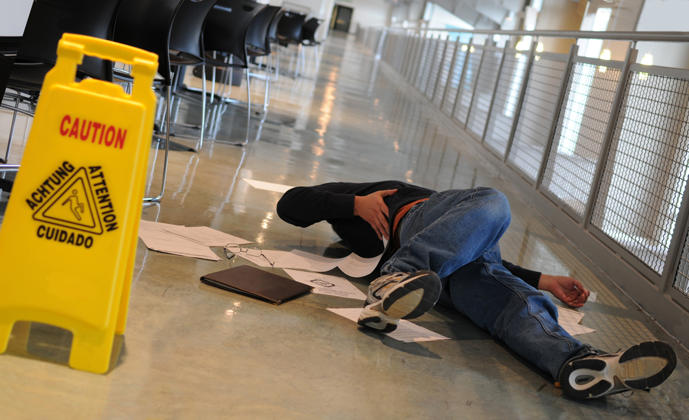 Image of a man laying on the ground with papers, a portfolio and glasses laying around him, a hand on his hip in pain. In the foreground, there's a yellow caution sign to warn against falling.