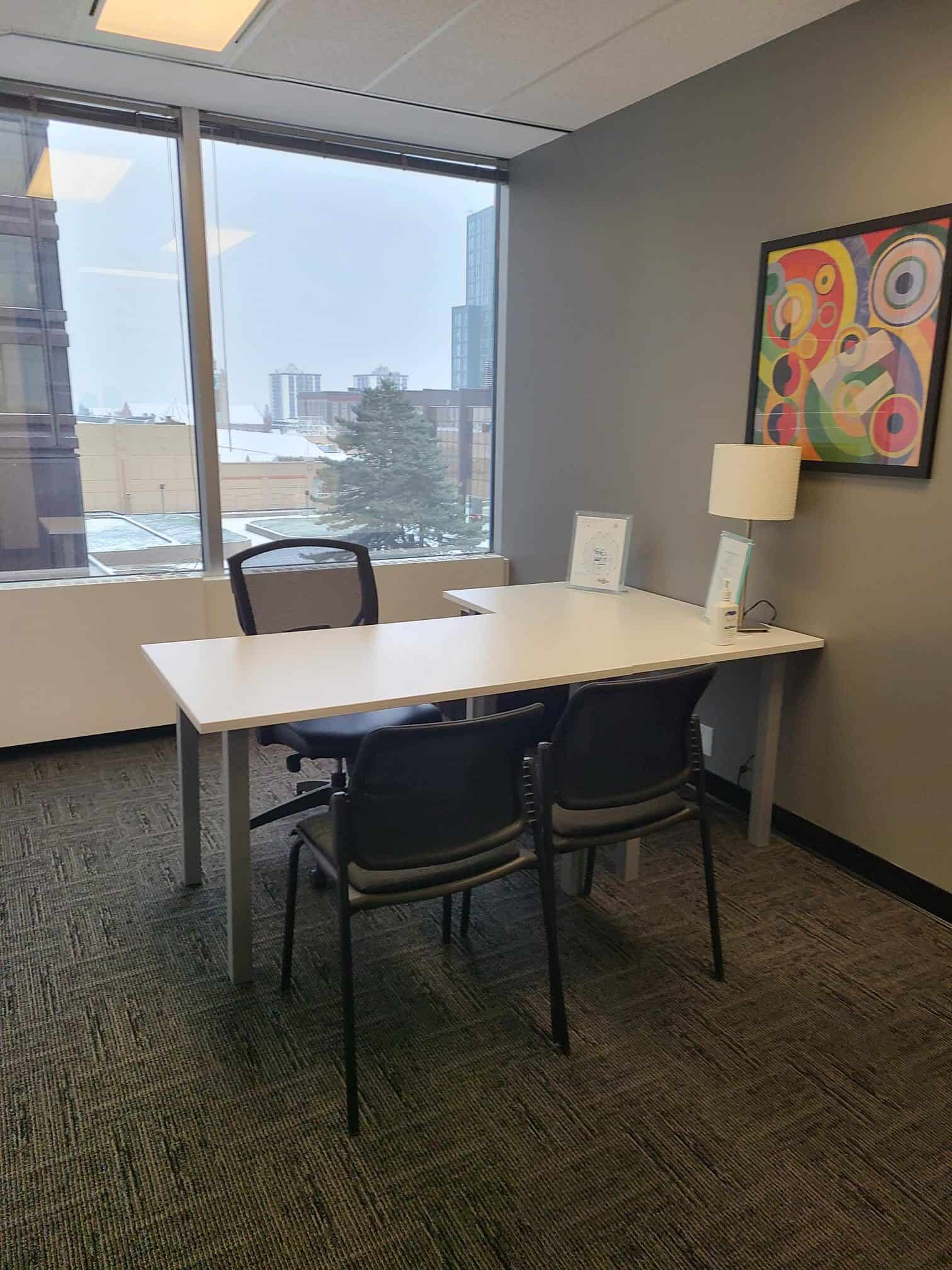 A picture of an office, with an L-shaped desk in front of a window with colorful art on the wall.