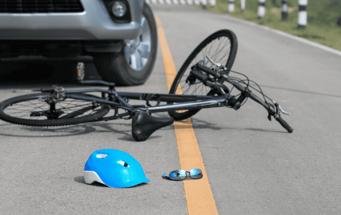 Image of a bike laying on a road after being struck by a car. There's also a blue helmet and sunglasses laying on the road.
